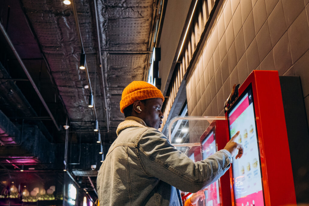 kiosk software Smiling African-American man in warm denim jacket with wireless earphones uses self-service kiosk to order snack in cafe